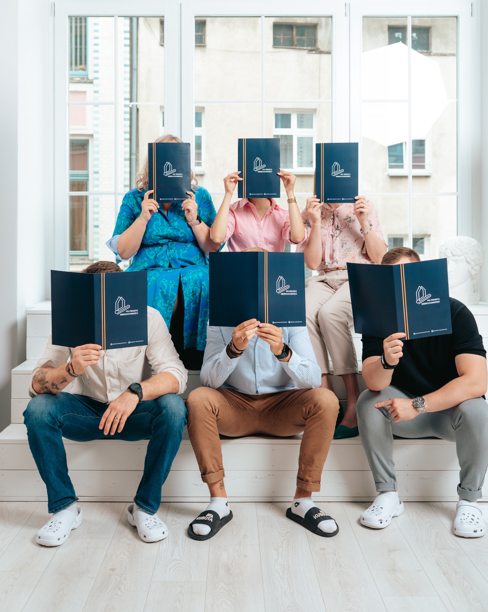 Zespół biura nieruchomości 'Po Prostu Nieruchomości' siedzi na schodach, zasłaniając twarze folderami z logo firmy na tle okna.