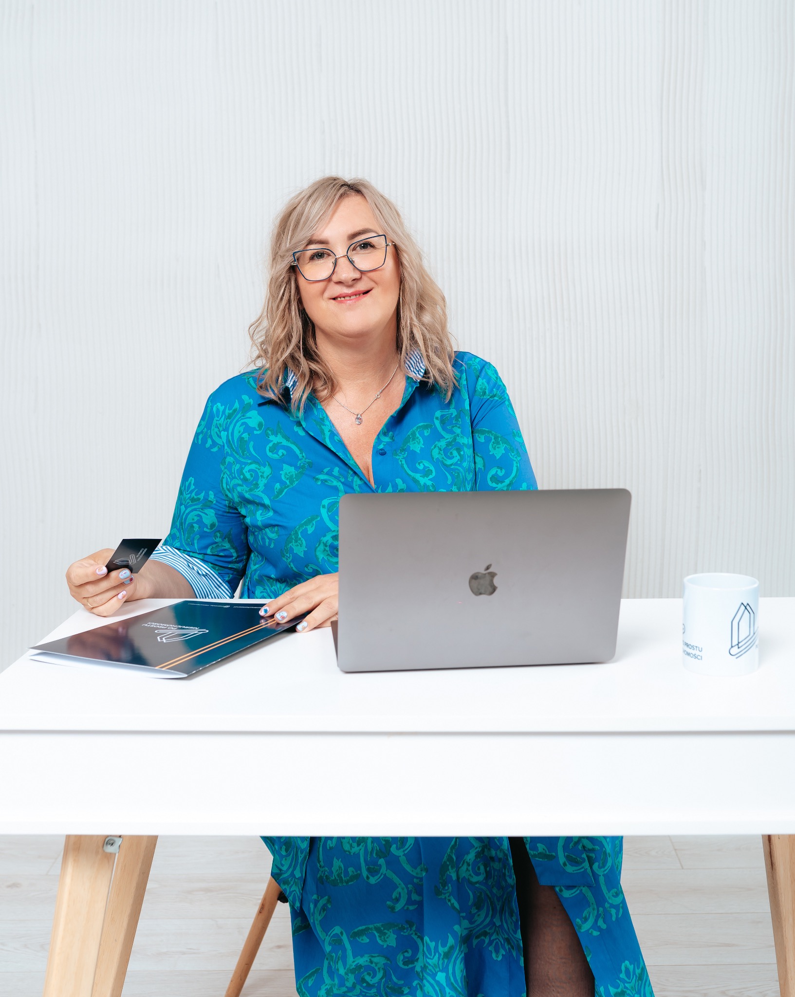 Kobieta w okularach siedzi przy biurku z laptopem, trzymając wizytówkę agencji nieruchomości i folder z logo firmy, obok stoi kubek z rysunkiem domu.
