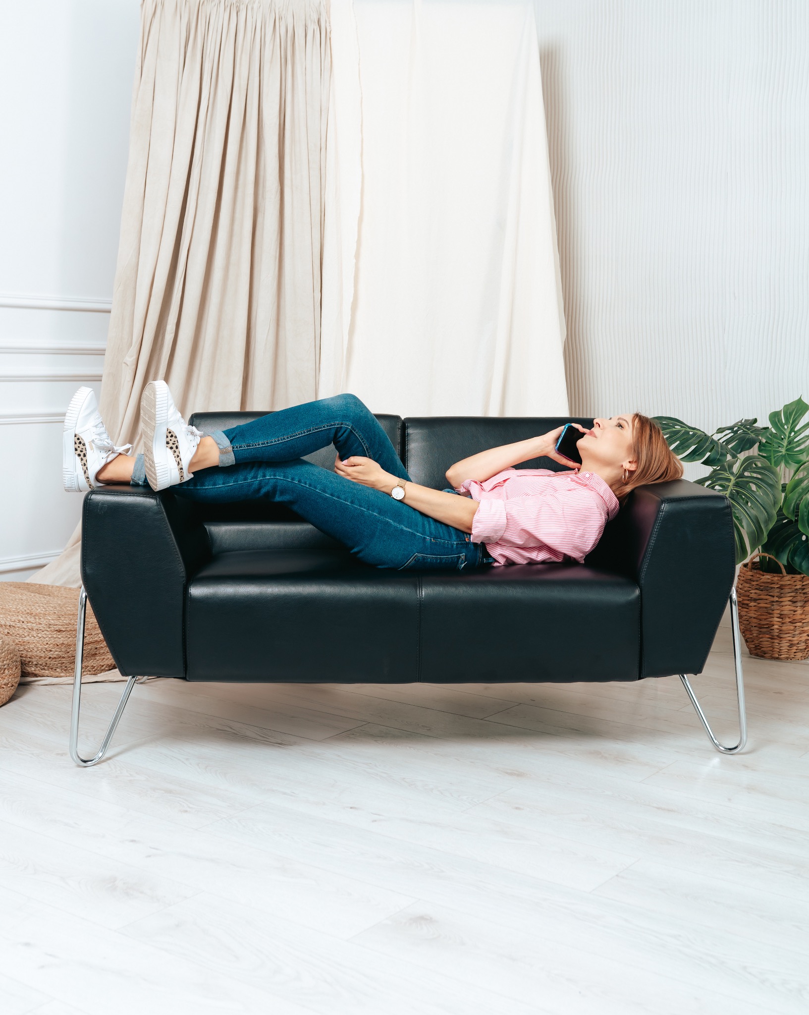 Kobieta w różowej koszuli i jeansach leży na czarnej skórzanej sofie, rozmawiając przez telefon w jasnym wnętrzu z rośliną doniczkową i beżowymi zasłonami.