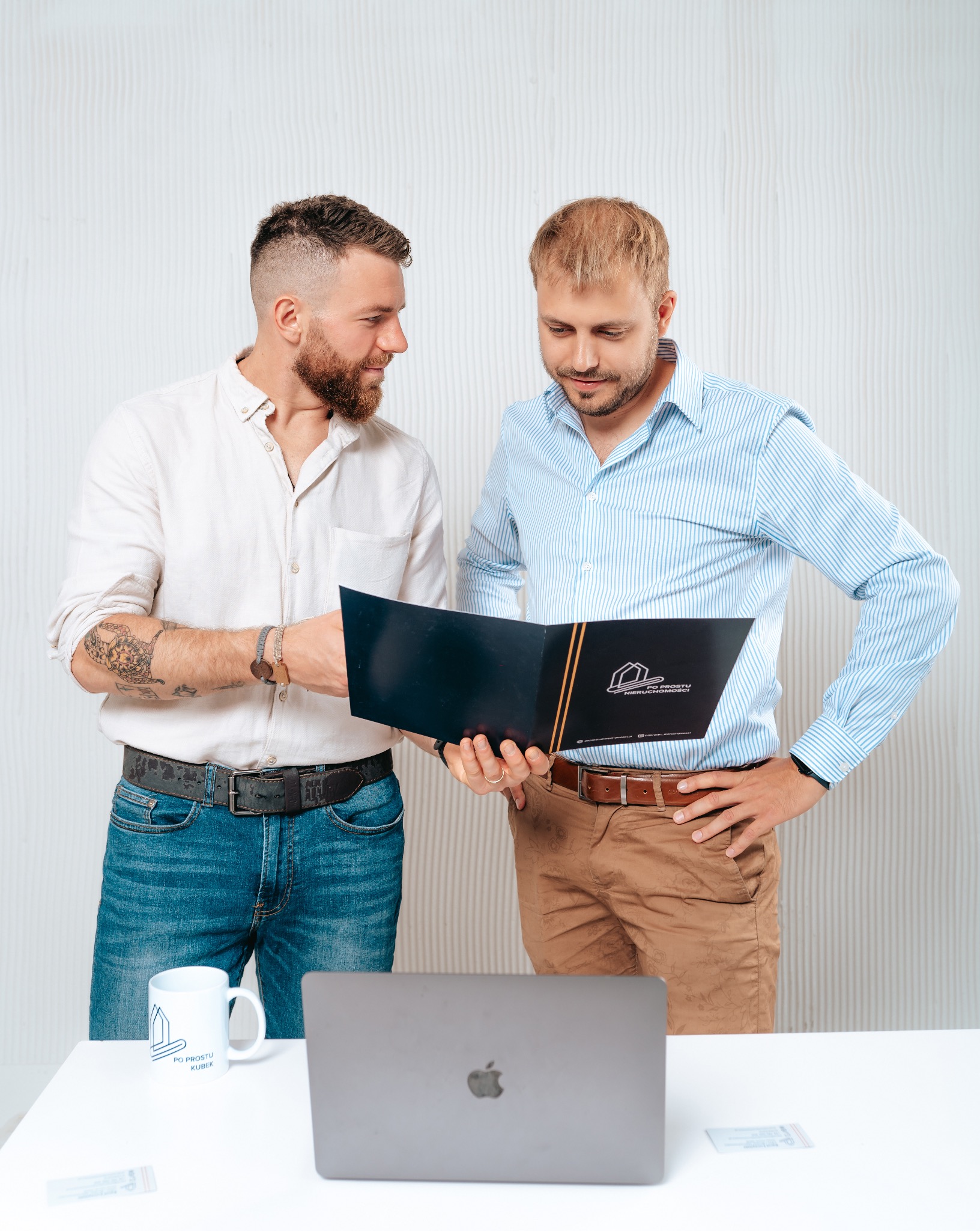 Dwóch mężczyzn w biurze analizuje dokument z logo firmy nieruchomości, widoczny laptop i firmowy kubek.