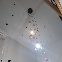 Ekoterm.plus - Biały sufit z punktowymi otworami i wiszącymi lampami w metalowych kloszach. Widoczny fragment drewnianej konstrukcji i zegar na ścianie.