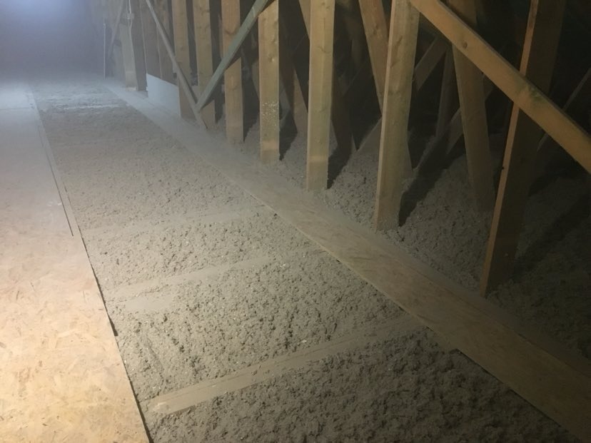 Niedokończona izolacja poddasza z widoczną wełną celulozową między drewnianymi krokwiami i deskami, oświetlona punktowo.