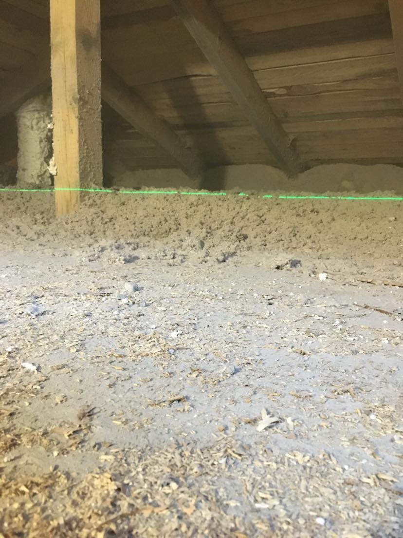 Poddasze z drewnianą konstrukcją dachu, częściowo wypełnione szarą izolacją, z widoczną zieloną linią poziomicy laserowej.