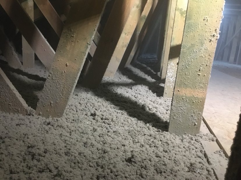 Wnętrze poddasza z drewnianą konstrukcją krokwiową wypełnione szarą, włóknistą izolacją, widoczne ślady pyłu na elementach drewnianych.