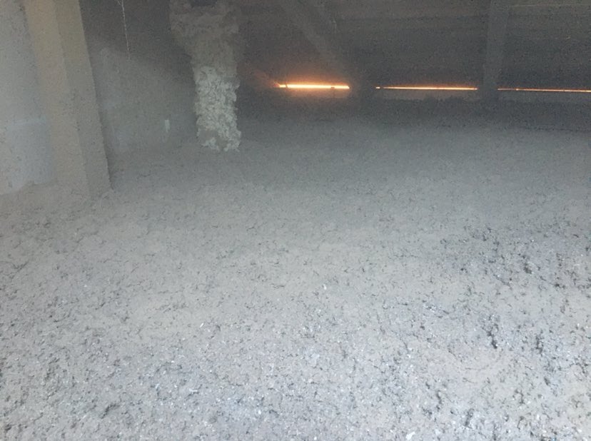 Szara, sypka izolacja termiczna pokrywająca podłogę poddasza, widoczne elementy konstrukcyjne dachu i fragment ściany.