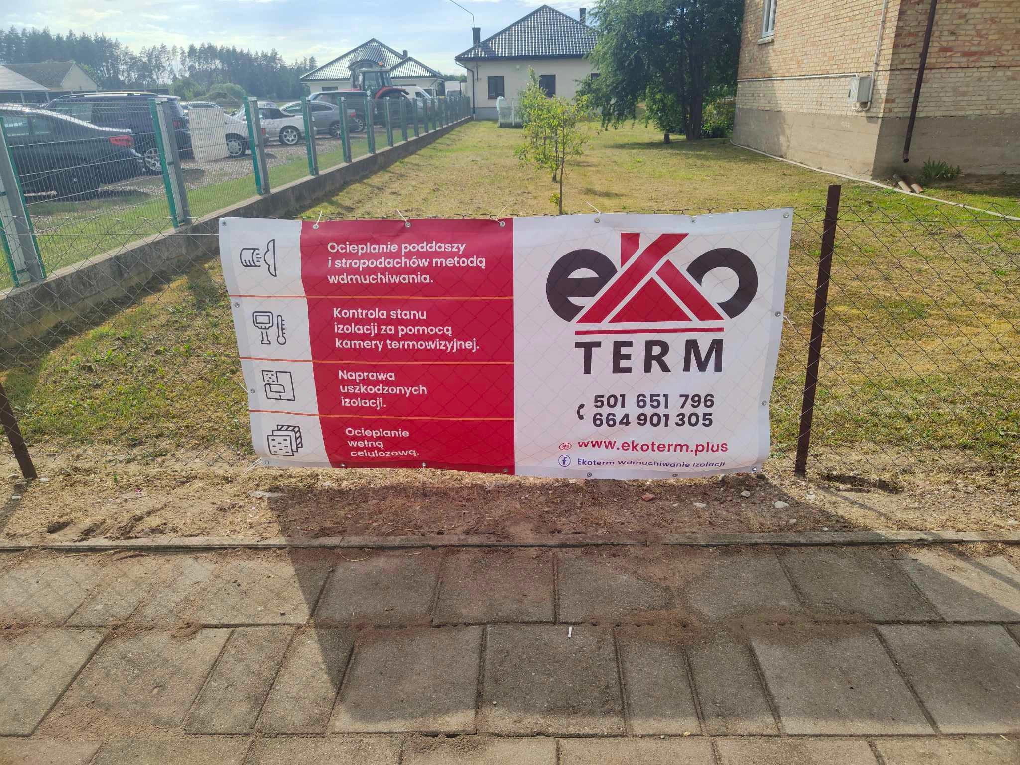Baner reklamowy firmy Eko-Term na trawniku, oferującej ocieplanie poddaszy metodą wdmuchiwania, kontrolę stanu izolacji kamerą termowizyjną i naprawę uszkodzonych izolacji, z widocznymi numerami...