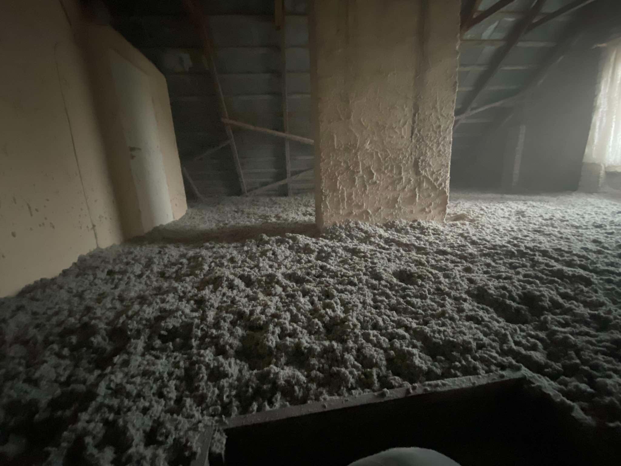 Niedokończone ocieplenie poddasza wełną mineralną, widoczne elementy konstrukcji dachu i komina, słabe oświetlenie.