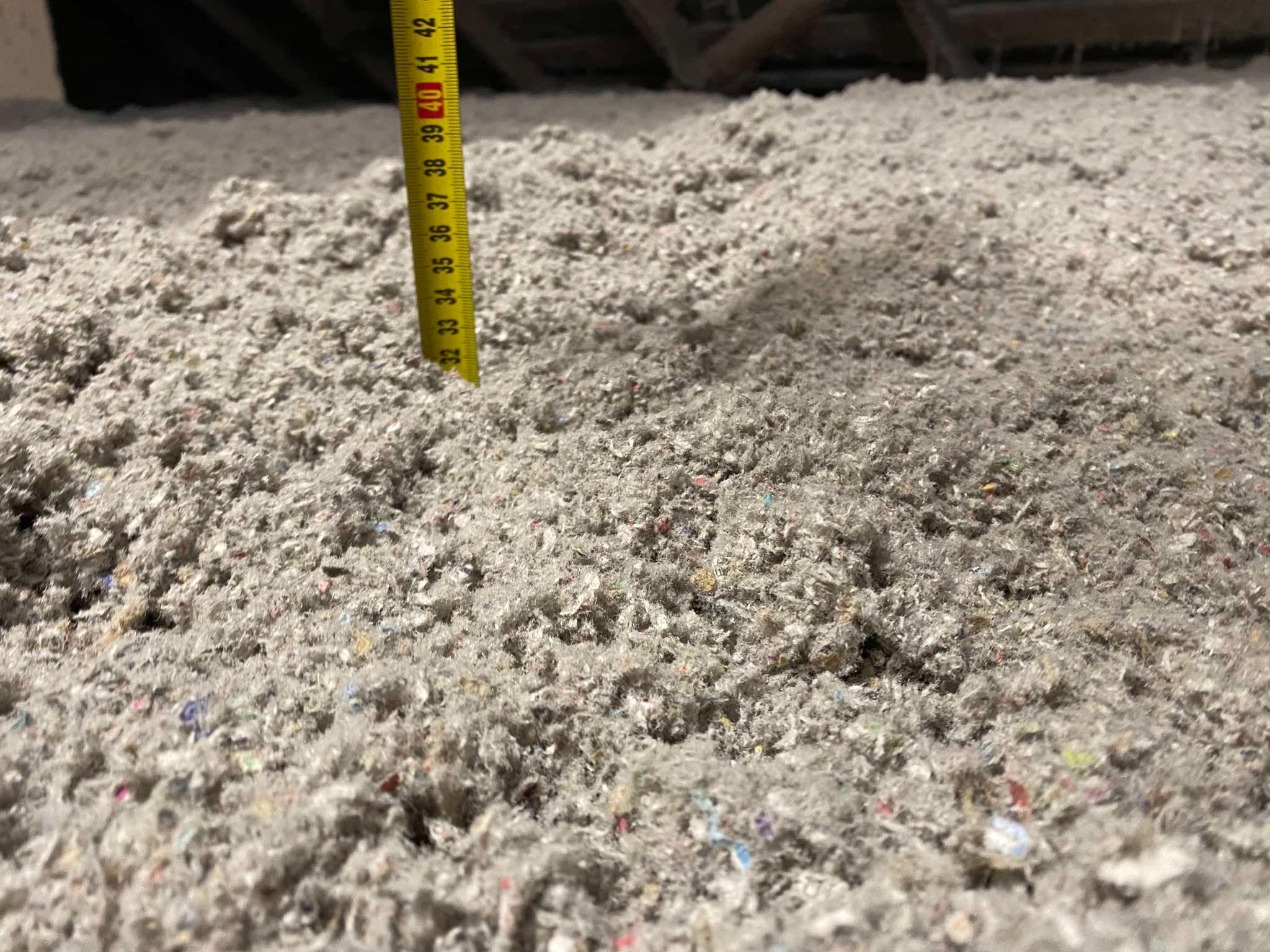 Luźno ułożona warstwa szarej wełny celulozowej na poddaszu, z żółtą miarką wskazującą grubość izolacji na około 40 cm.