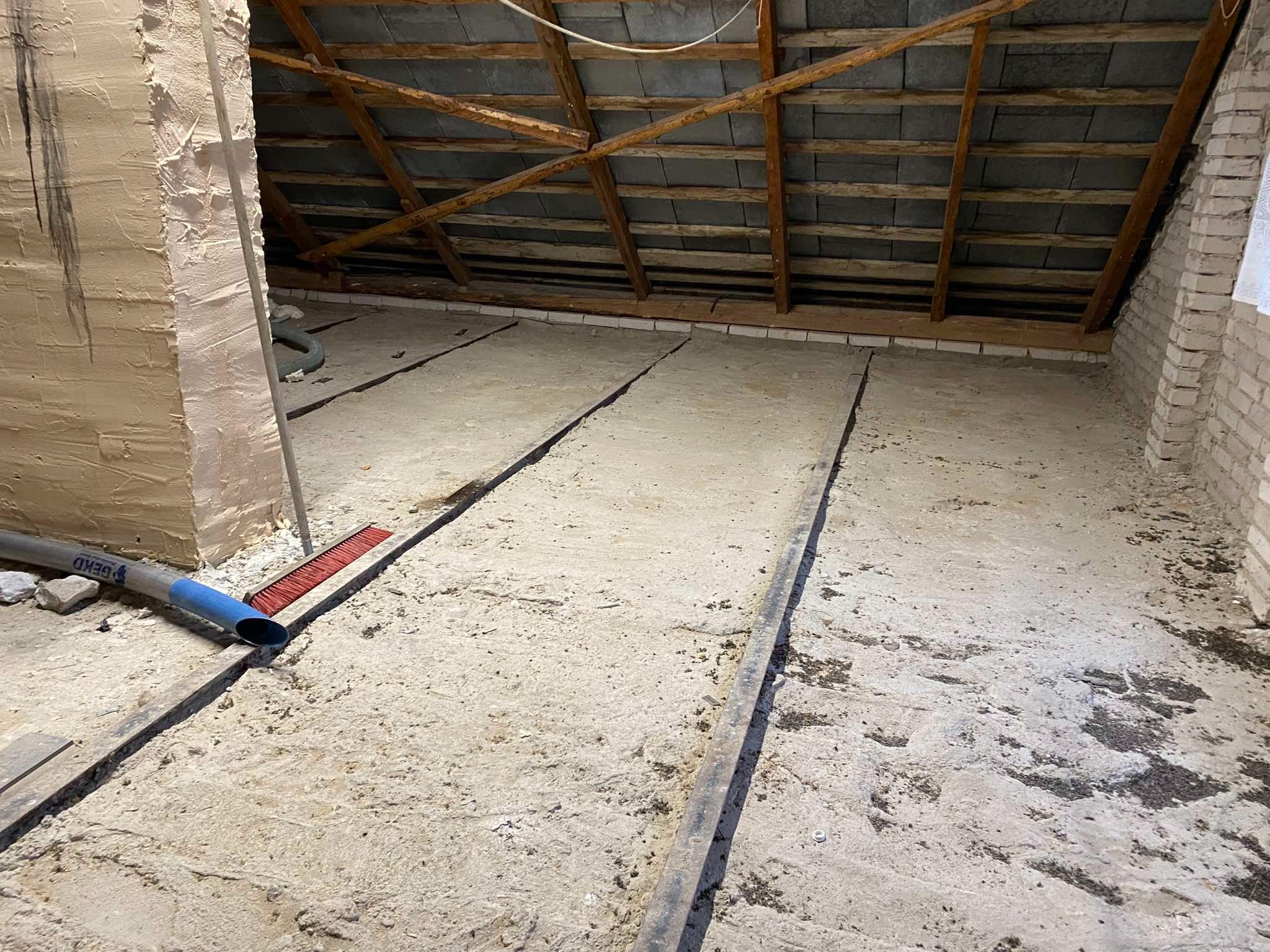 Przygotowanie podłoża na poddaszu: szlichta z wyznaczonymi dylatacjami, widoczny fragment więźby dachowej, ściana z surowej cegły oraz narzędzia pracy - szczotka i rura od odkurzacza przemysłowego.