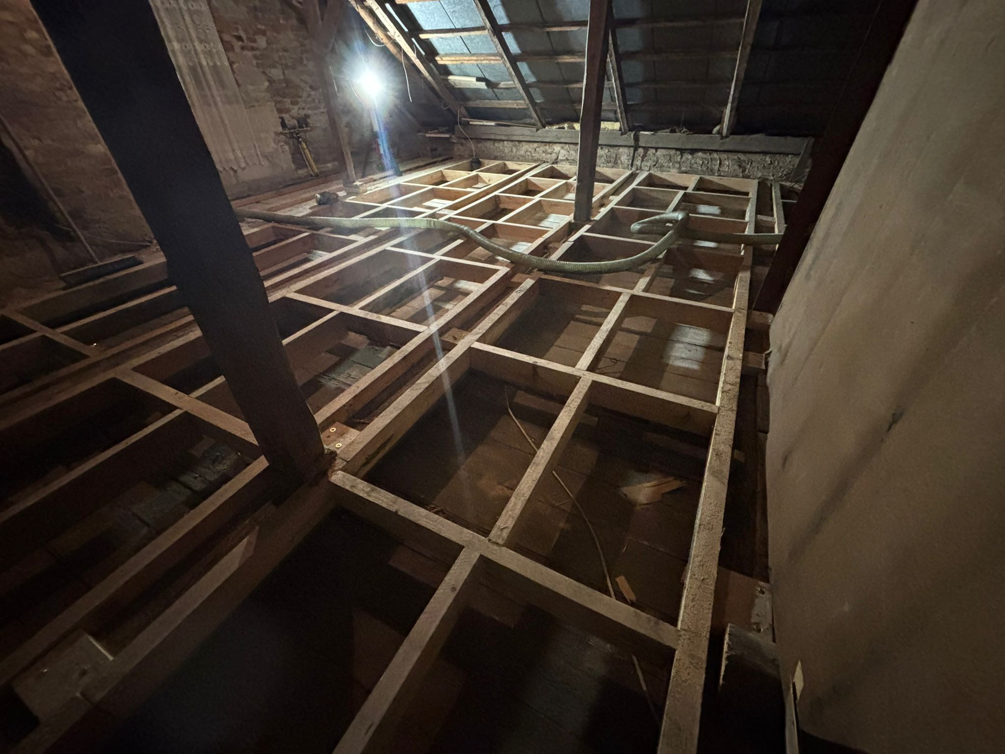 Poddasze z drewnianą konstrukcją szkieletową przygotowaną do ocieplenia, widoczny wąż do wdmuchiwania izolacji. Nierówne oświetlenie podkreśla surowość wnętrza.