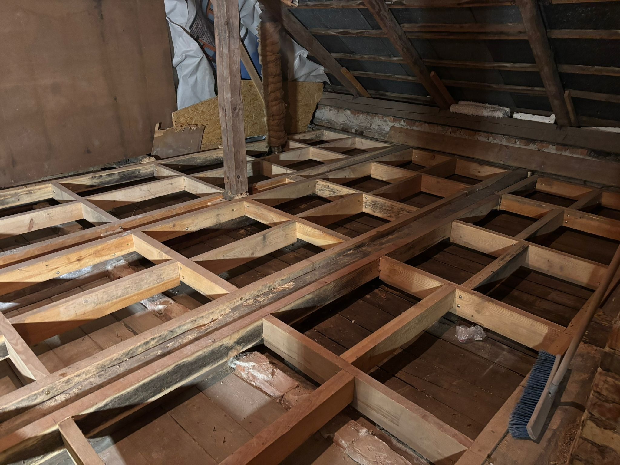 Drewniana konstrukcja podłogi na poddaszu, przygotowana do izolacji. Widoczne stare deski podłogowe i fragment dachu. Ujęcie z góry, perspektywa wnętrza.
