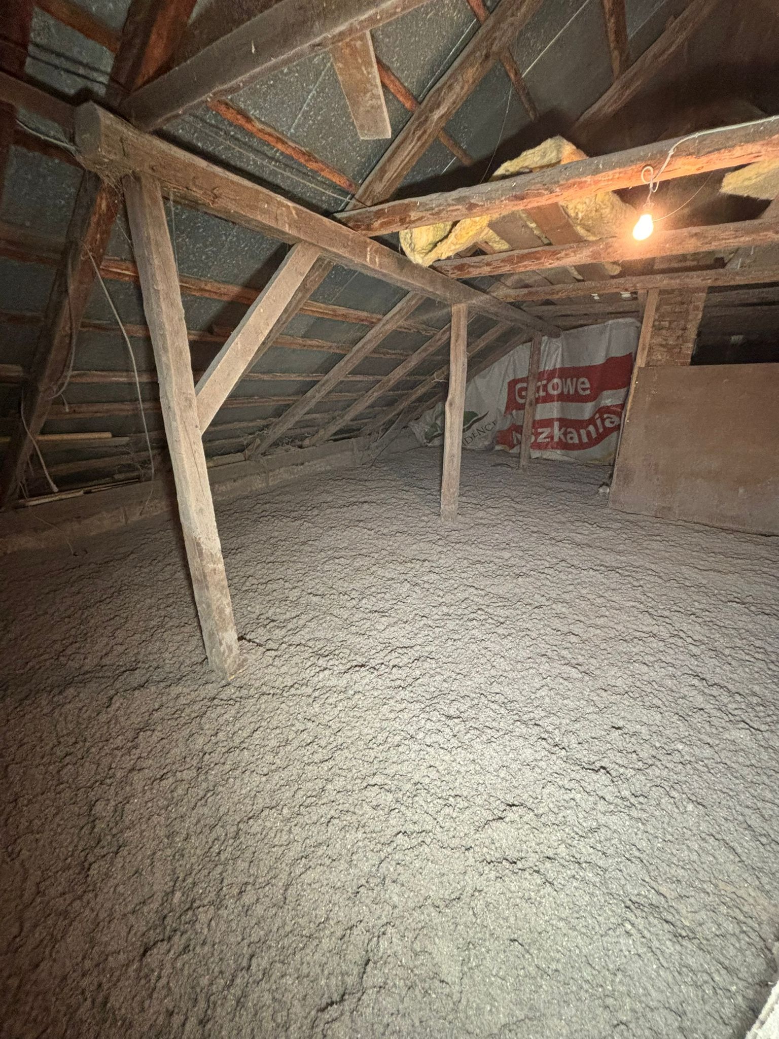 Poddasze po izolacji, widoczne drewniane elementy konstrukcyjne i warstwa szarej izolacji. W tle fragment reklamy materiałów izolacyjnych.