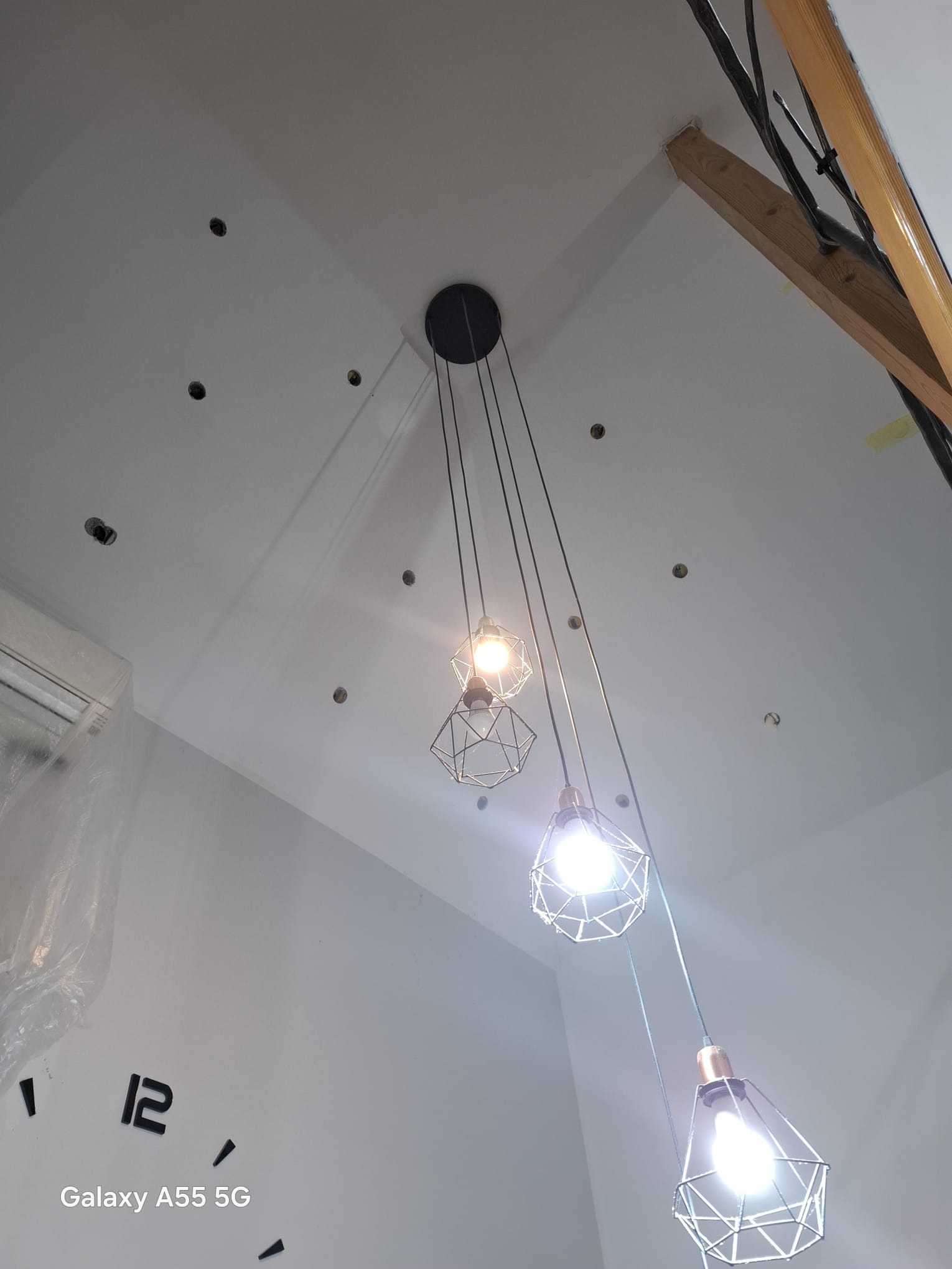 Biały sufit z punktowymi otworami i wiszącymi lampami w metalowych kloszach. Widoczny fragment drewnianej konstrukcji i zegar na ścianie.