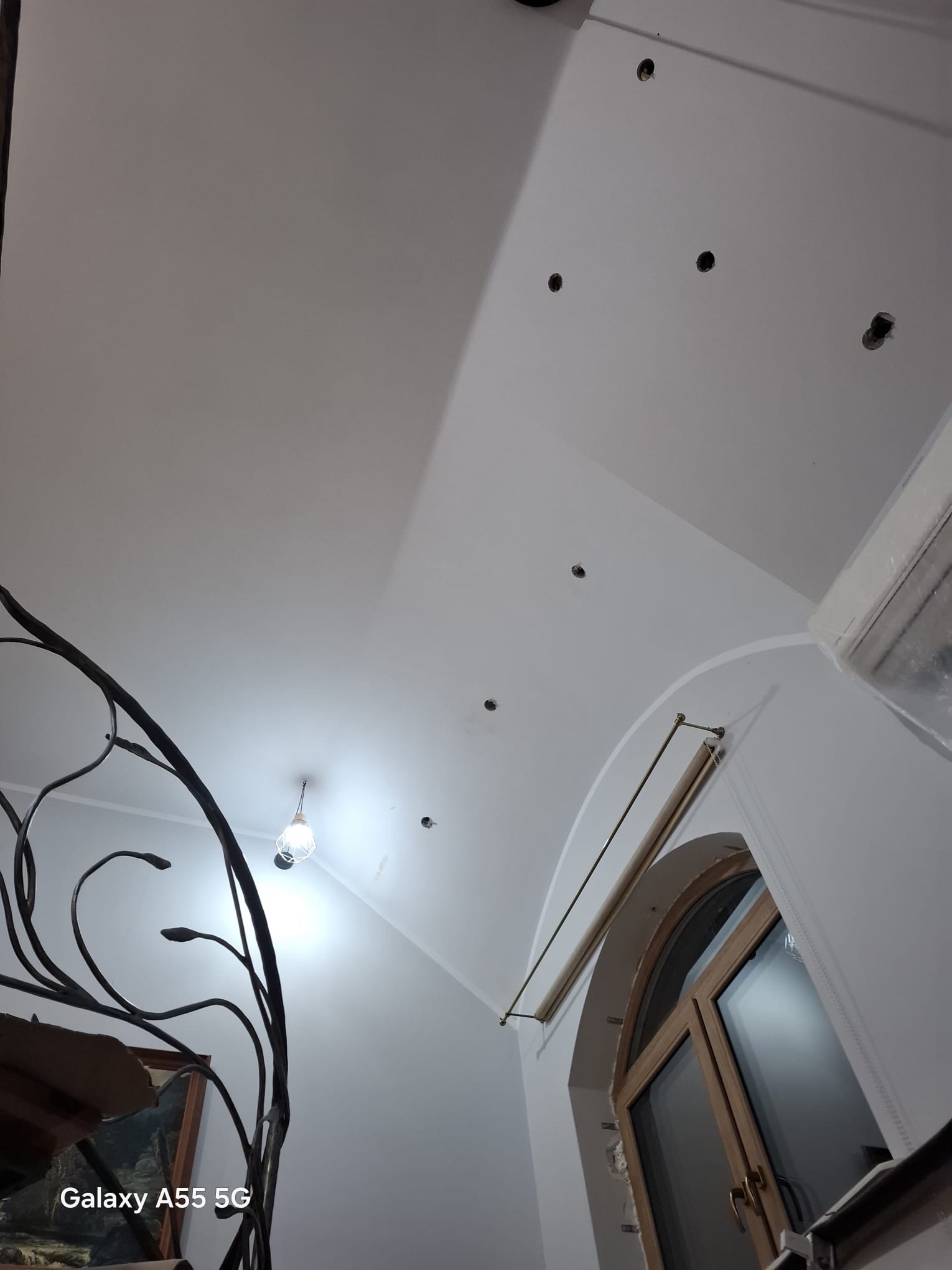 Białe poddasze z widocznymi otworami na oświetlenie, fragmentem okna i ozdobną, metalową balustradą schodów. Widok z dołu, w trakcie prac wykończeniowych.