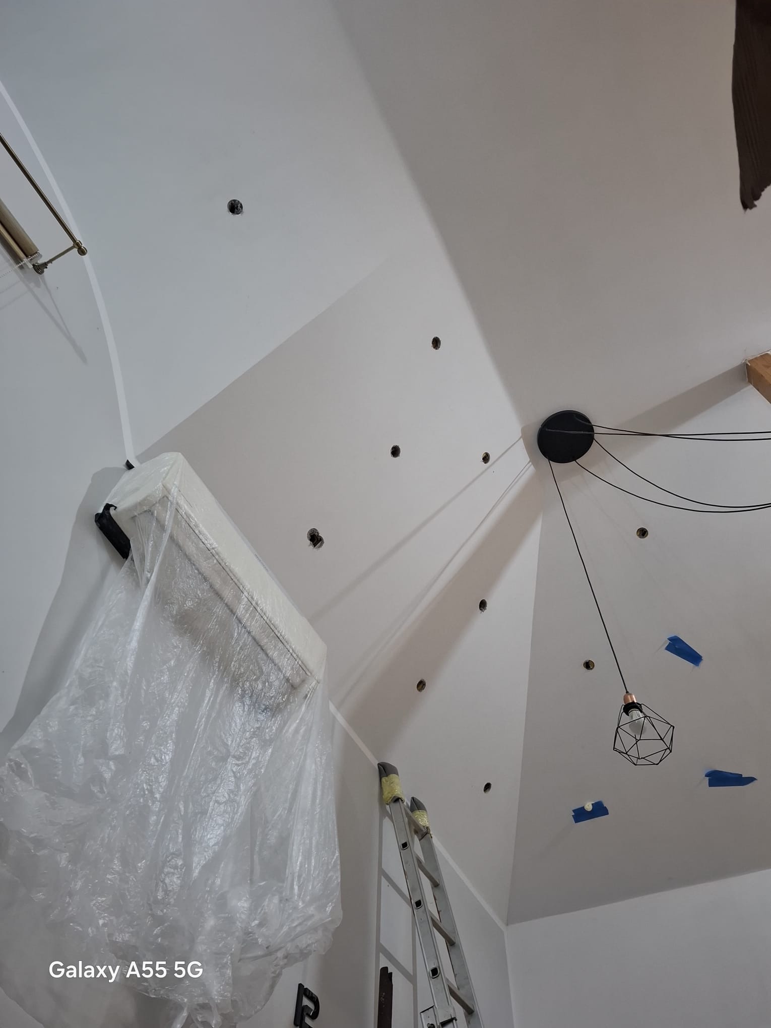 Poddasze po przygotowaniu do prac: zabezpieczony klimatyzator folią, drabina, otwory w suficie, lampa wisząca, niebieska taśma malarska. Białe ściany i sufit.