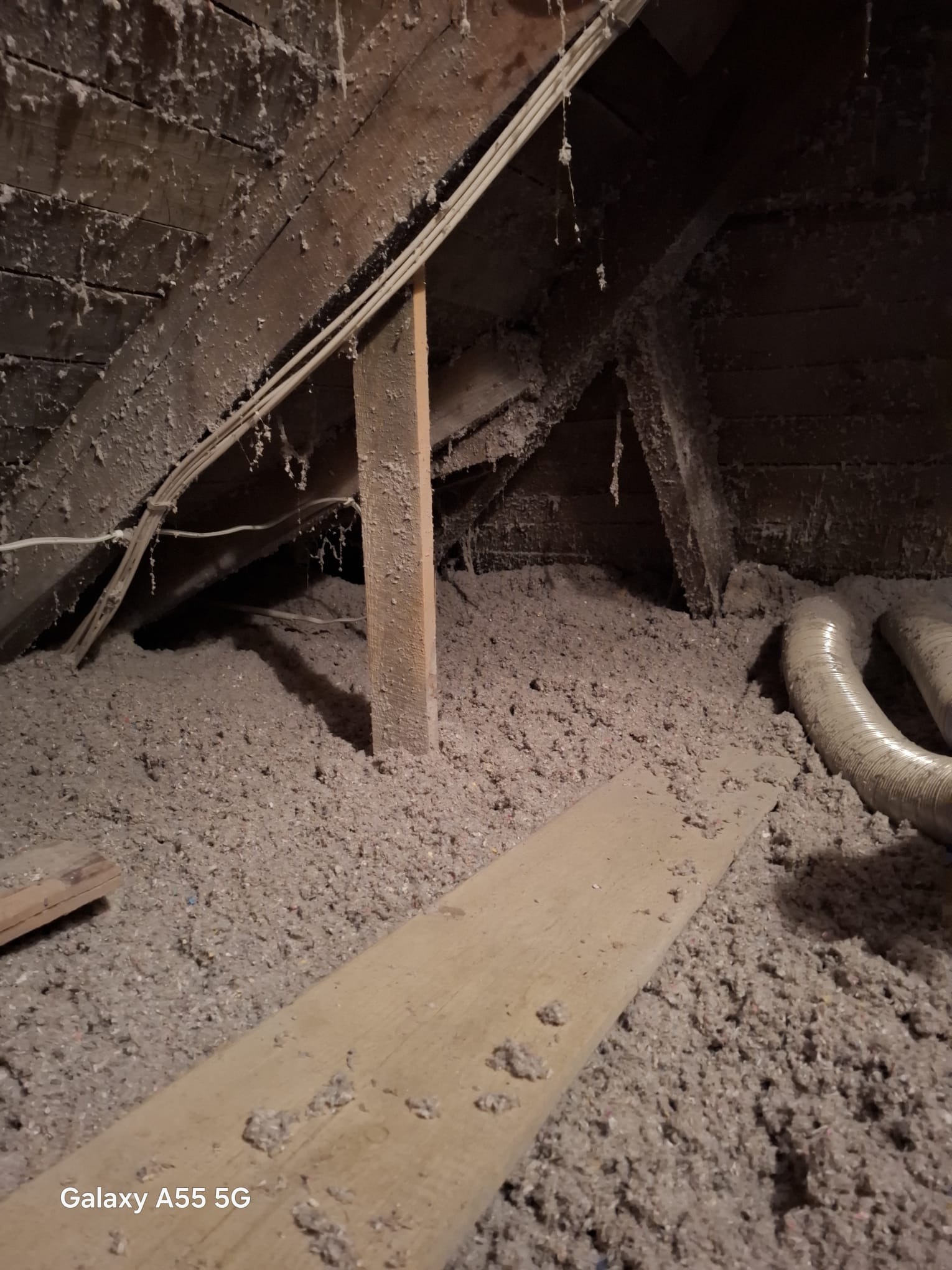 Poddasze z widoczną warstwą izolacji z wełny mineralnej, drewnianą konstrukcją dachu i fragmentem rury wentylacyjnej. Widoczne niedoskonałości i zabrudzenia.