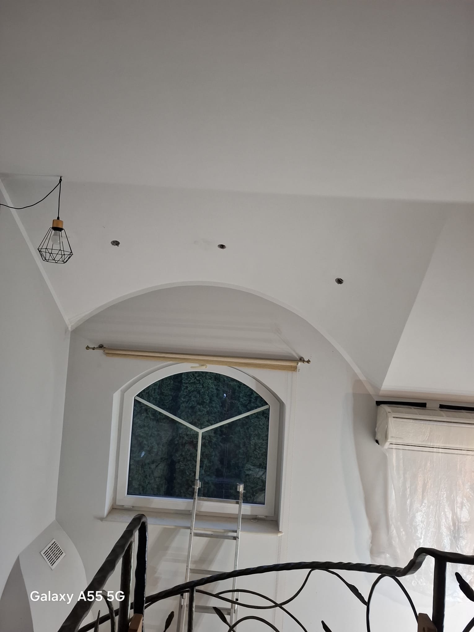 Poddasze po remoncie: białe ściany, okno z łukiem, metalowe schody, lampa wisząca. Widoczne ślady po montażu oświetlenia sufitowego. Klimatyzator zabezpieczony folią.