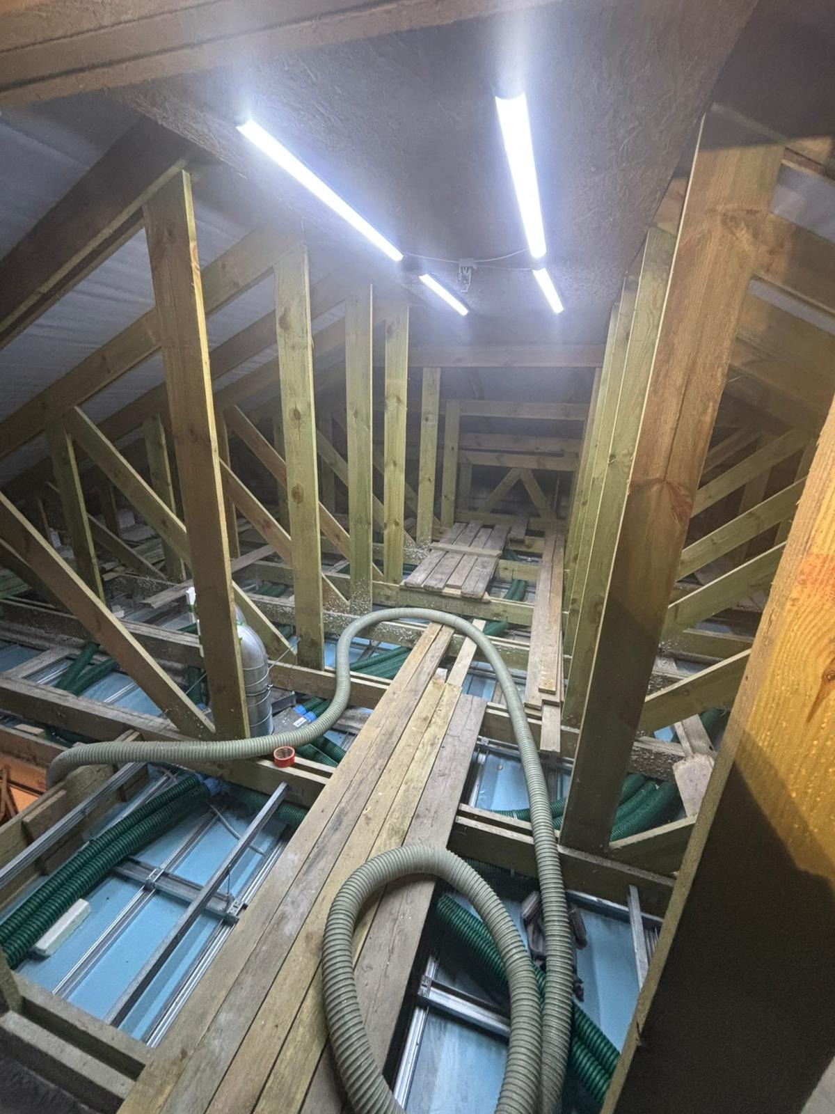 Poddasze w trakcie prac: widoczne drewniane krokwie, zielona instalacja wentylacyjna i szare rury. Oświetlenie jarzeniowe. Fragment podłogi z desek.