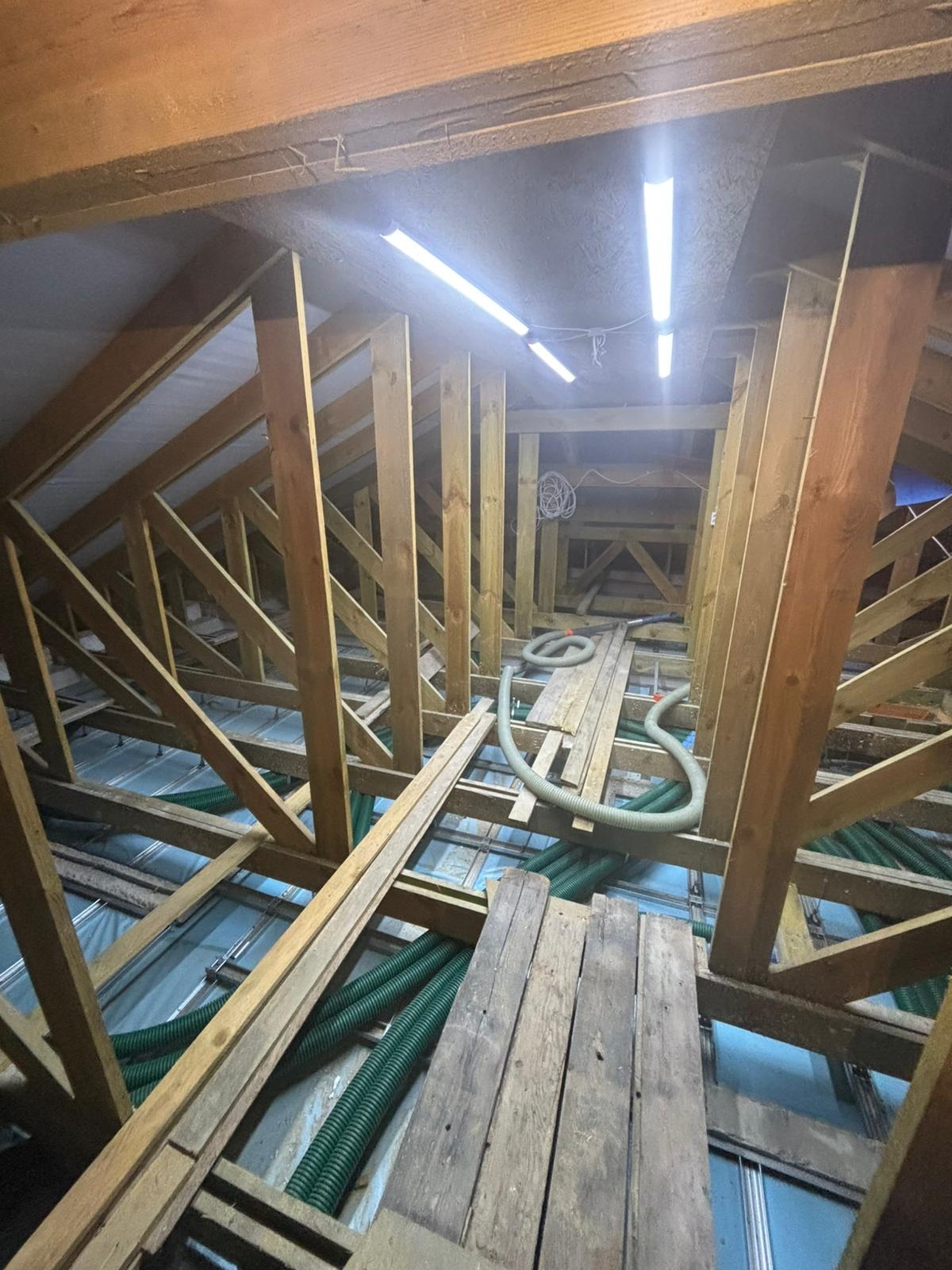 Skomplikowana konstrukcja drewniana poddasza z widocznymi elementami instalacji wentylacyjnej w postaci zielonych rur. Widoczne deski i oświetlenie.