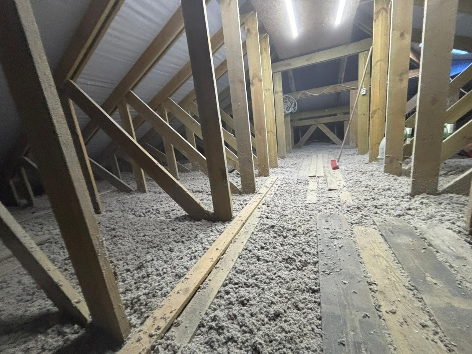 Poddasze z widoczną izolacją z wełny mineralnej, drewnianą konstrukcją więźby dachowej i fragmentami desek podłogowych. Widoczne oświetlenie i elementy instalacji.