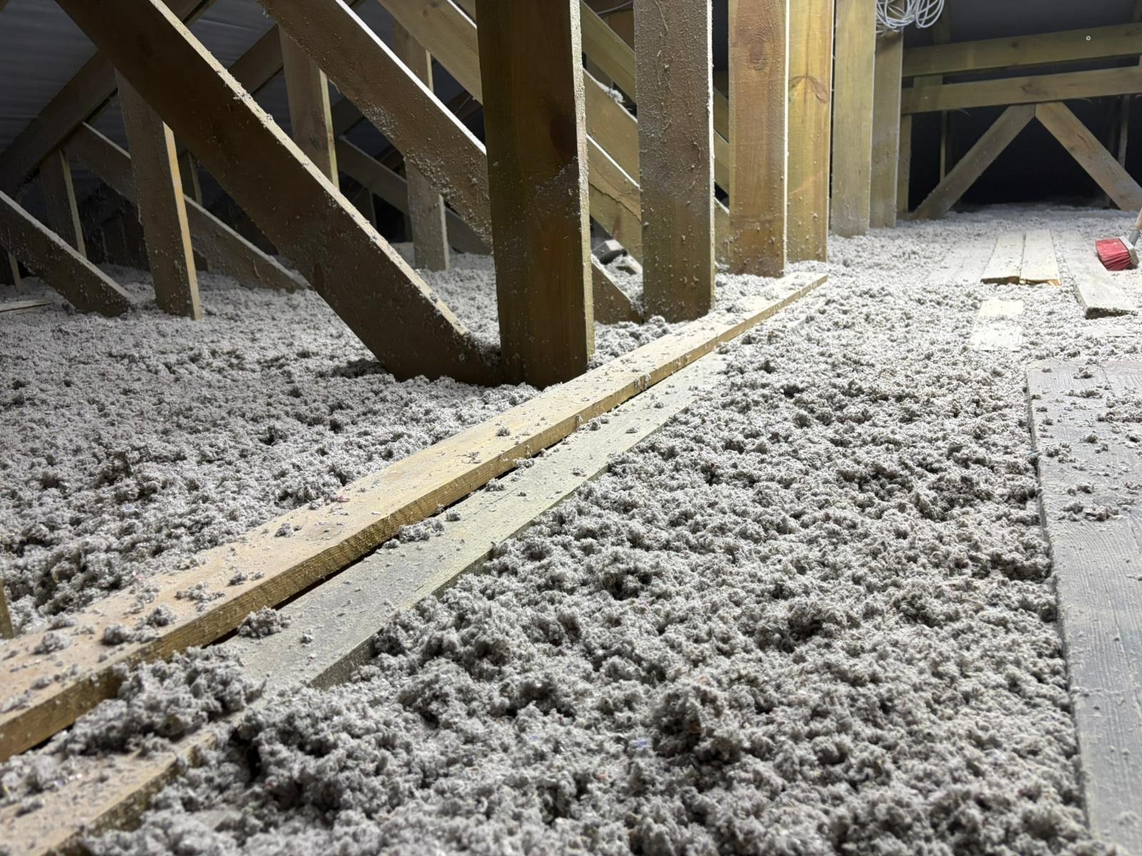 Poddasze z drewnianą konstrukcją wypełnione szarą izolacją celulozową, widoczne deski i fragment instalacji elektrycznej.