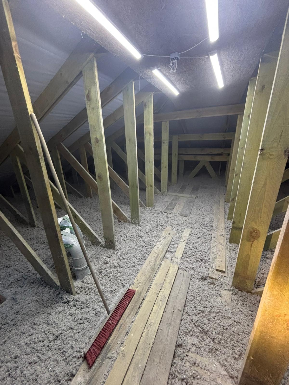 Poddasze z drewnianą konstrukcją wypełnione szarą izolacją. Widoczne deski, rura wentylacyjna i szczotka. Oświetlenie jarzeniowe.