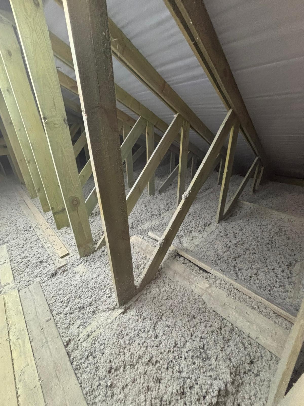 Skośny kadr na poddasze z drewnianą konstrukcją i szarą warstwą izolacyjną. Widoczne fragmenty desek podłogowych i biała membrana dachowa.