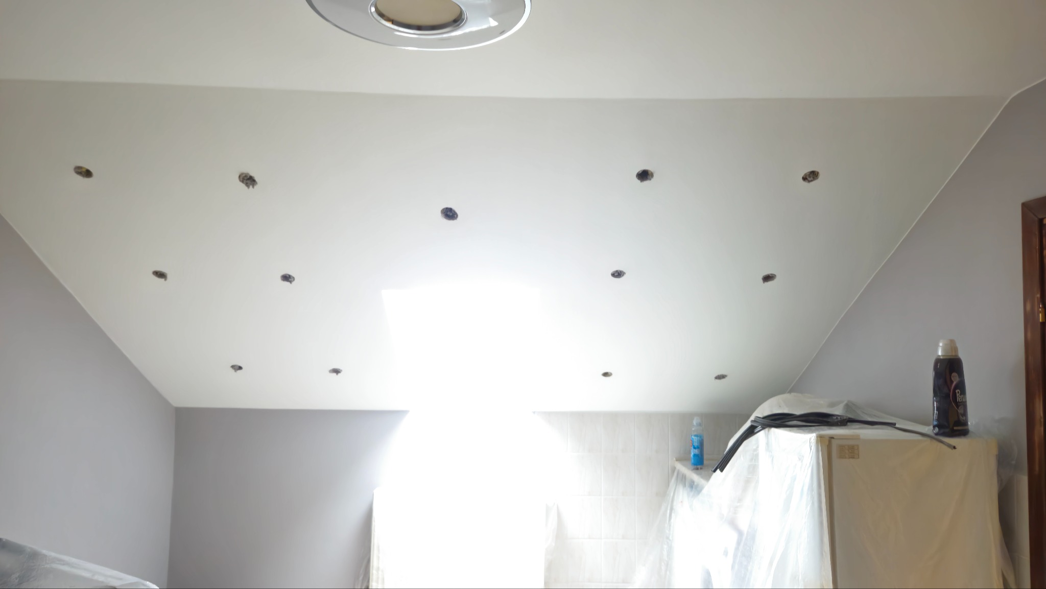 Białe poddasze ze skośnym sufitem, przygotowane do montażu oświetlenia punktowego. Widoczne otwory w suficie i zabezpieczone meble.