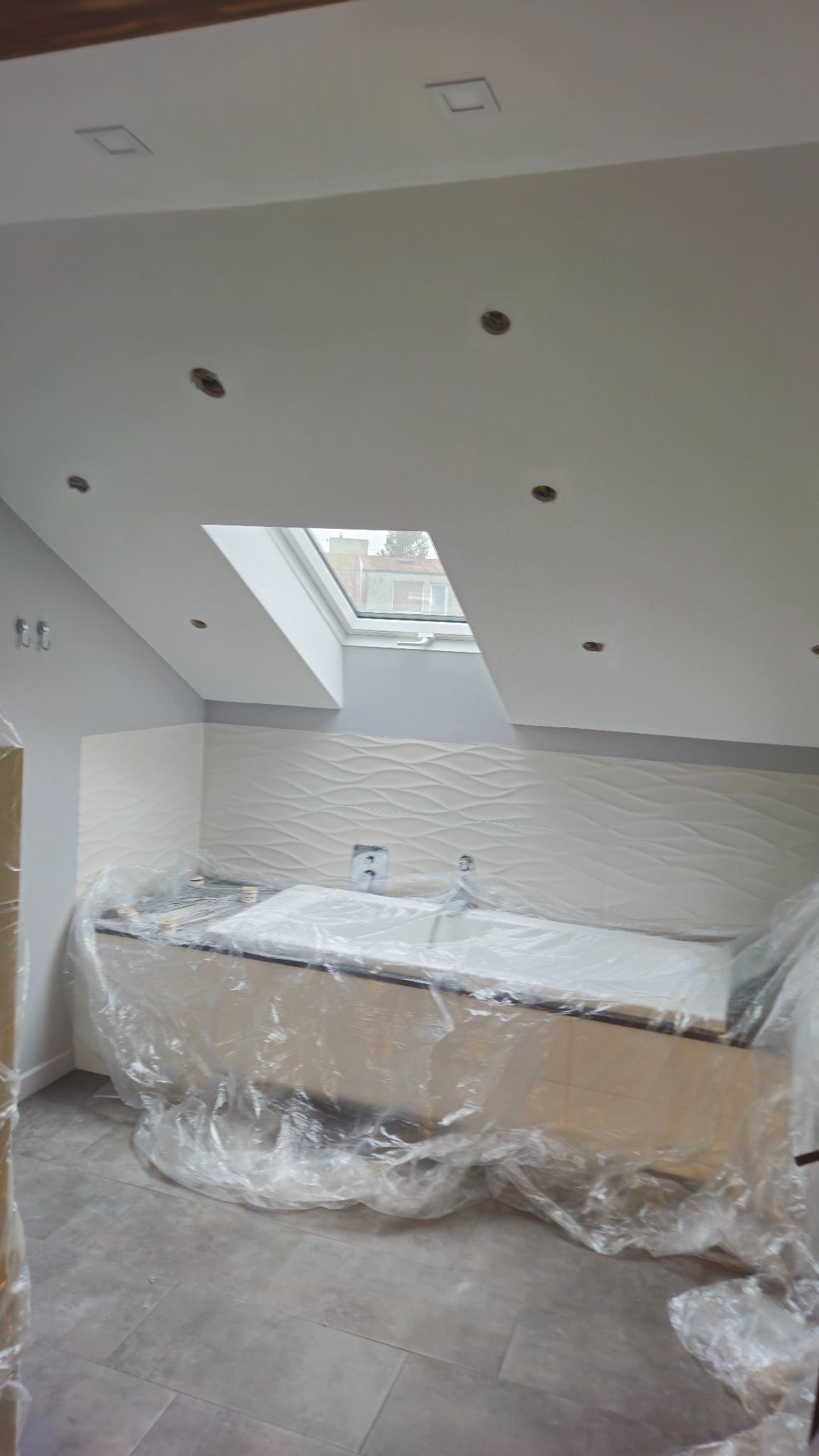 Poddasze z oknem dachowym i wykończoną ścianą z dekoracyjną płytką, przygotowane do dalszych prac wykończeniowych, zabezpieczone folią ochronną.