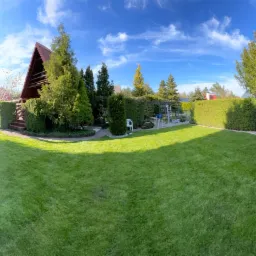 Panorama zadbanego ogrodu z równo przystrzyżonym trawnikiem, żywopłotem i drzewami iglastymi, obrzeża rabat wysypane białym kamieniem, w tle domek letniskowy z drewnianymi schodami.