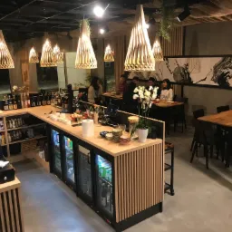 Wnętrze restauracji z drewnianym barem, wiszącymi lampami o ażurowym designie i klientami przy stolikach. Uwagę zwraca falujący sufit i grafika na ścianie.