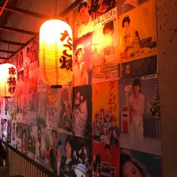 Wnętrze lokalu gastronomicznego z dekoracją ściany w postaci kolażu plakatów w stylu japońskim, oświetlone wiszącymi lampionami z napisami w języku japońskim.