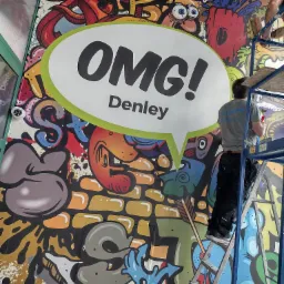 Mural z kolorowymi graffiti i napisem 'OMG! Denley' w dymku, malowany przez mężczyznę stojącego na rusztowaniu.