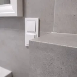 Szara, matowa płytka ceramiczna na ścianie łazienki, z widocznym narożnikiem obudowy i białym włącznikiem światła oraz fragmentem białej ramy okna w górnym rogu.