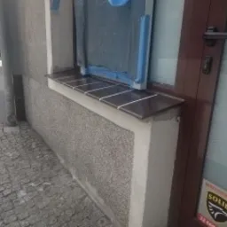 Zamontowany parapet zewnętrzny z ciemnobrązowych płytek ceramicznych, z widocznymi fugami, na tle szarej, chropowatej elewacji i częściowo zasłoniętego okna z brązową ramą.