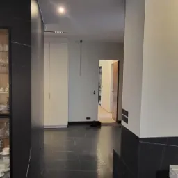 Wnętrze mieszkania z czarną marmurową podłogą i ścianami, widok na korytarz z białymi szafami i drzwiami.