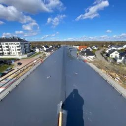 Długi, szary dach z blachy na nowym budynku, widok z góry z cieniem fotografa, w tle zabudowa miejska i błękitne niebo z chmurami.