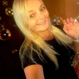 Uśmiechnięta kobieta z blond włosami dmucha bańki mydlane, trzymając dłoń w powietrzu, na ciemnym tle.