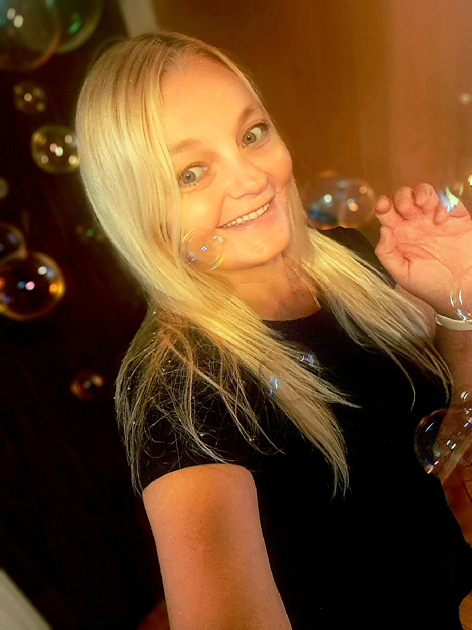 Uśmiechnięta kobieta z blond włosami dmucha bańki mydlane, trzymając dłoń w powietrzu, na ciemnym tle.