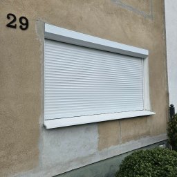 Pentra Plus - Biała roleta zewnętrzna na oknie budynku z numerem 29, widoczna z poziomu gruntu. Roleta opuszczona, zasłania okno. Tynk w odcieniach beżu i szarości.