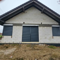 Pentra Plus - Surowe ściany budynku z trzema oknami zabezpieczonymi czarnymi roletami zewnętrznymi, widok z zewnątrz na tle pochmurnego nieba i częściowo zagospodarowanego terenu.