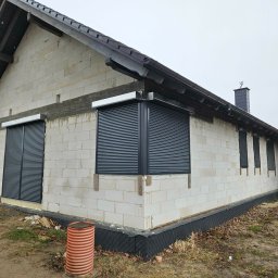 Pentra Plus - Dom w trakcie budowy z zamontowanymi, grafitowymi roletami zewnętrznymi na oknach. Widoczna surowa elewacja z betonu komórkowego i czarne orynnowanie.
