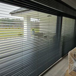 Pentra Plus - Ciemne żaluzje poziome zamontowane w oknie, widok na zewnątrz z zielenią i nowoczesną architekturą. Częściowo zasłonięte meble w folii ochronnej.