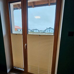 Pentra Plus - Drewniane okno z beżowymi plisowanymi roletami wewnętrznymi, widok na domy za oknem. Plisy częściowo zasłonięte, widoczna faktura materiału i mechanizm regulacji.