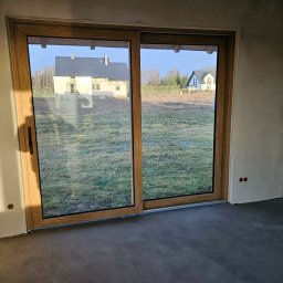 Pentra Plus - Przesuwne okno tarasowe z drewnianą ramą, widok na zewnątrz z dwoma domami w tle. Surowe ściany wewnątrz, wylewka podłogowa.