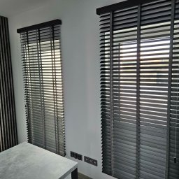 Pentra Plus - Dwa okna z drewnianymi żaluzjami w odcieniach szarości, z widocznymi sznurkami do regulacji. Obok dekoracyjny panel ścienny z pionowych listew w podobnej kolorystyce.