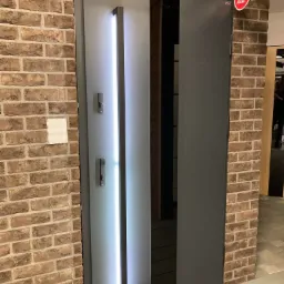 Ciemnoszare drzwi wejściowe z podświetlanym panelem pionowym, zamontowane w murze z cegły, z widocznym fragmentem ekspozycji w salonie.