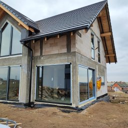 Pentra Plus - Dom w trakcie budowy z zamontowanymi dużymi oknami przesuwnymi i trójkątnym oknem na szczycie, widoczne materiały budowlane i inne domy w tle.