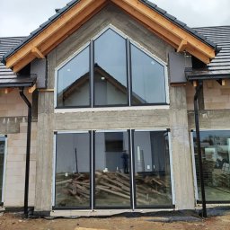 Pentra Plus - Surowy stan budynku z zamontowanymi oknami o czarnych ramach, widoczne drewniane elementy konstrukcji dachu oraz porozrzucane deski na zewnątrz.