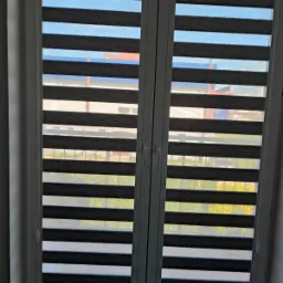 Dwuskrzydłowe okno PCV z zamontowanymi roletami dzień noc w odcieniach szarości, widok z wnętrza pomieszczenia na zewnątrz z zarysem zabudowy miejskiej i zieleni.